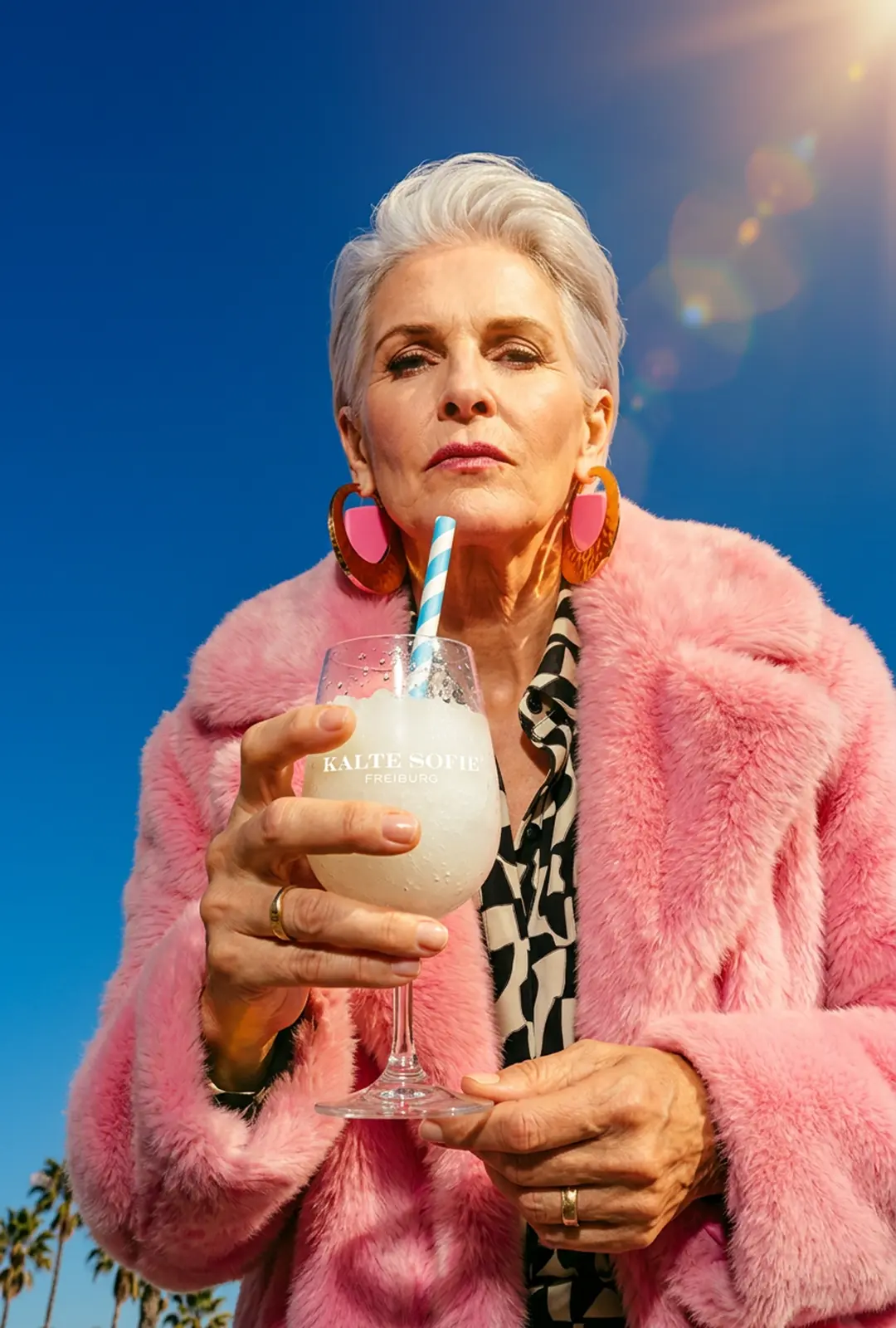 Loretta hält eine eisgekühlte Kalte Sofie Freiburg Glas mit Strohhalm in der Hand, trägt einen pinkfarbenen Pelzmantel – stilvoll und selbstbewusst vor blauem Himmel.